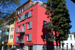 Rot gestrichenes viergeschossiges Mehrfamilienhaus; Holzaufsatz auf Dachterrasse mit Sonnenschirm; graue Verschattungselemente an den Fenstern