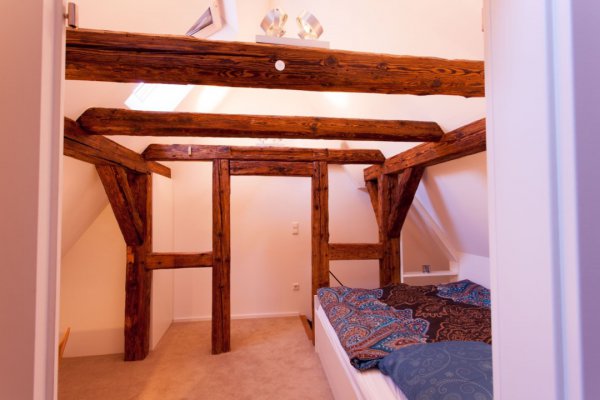 Dachbodenraum, in dem die Holzbalken-Ständer gestrichen noch sichtbar sind; rechts ein Bett, Wände weiß gestrichen.