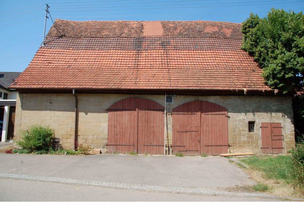 Längliches Scheunen-Gebäude von Traufseite mit älterem Dach und zwei rötlich-braunen Toren im Erdgeschoss